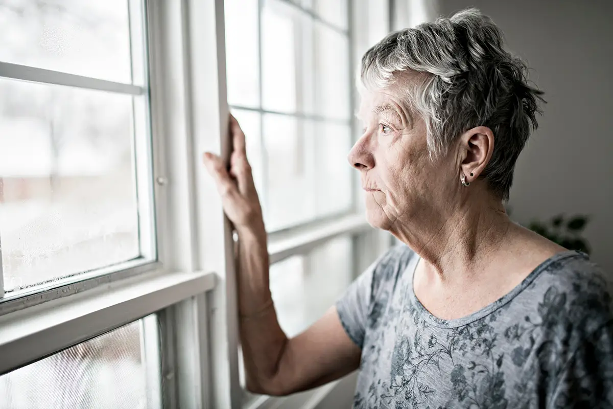 Photo of female senior looking somberly out of the window of her home