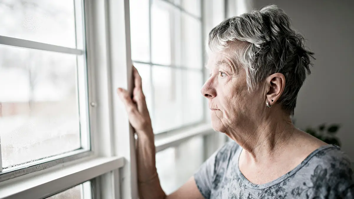 Photo of female senior looking somberly out of the window of her home