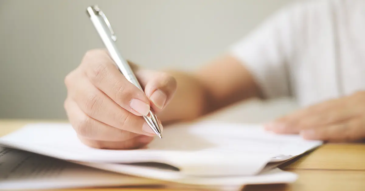 Photo of a person filling out a paper application on a desk with a pen. Photo by Adobe Stock.