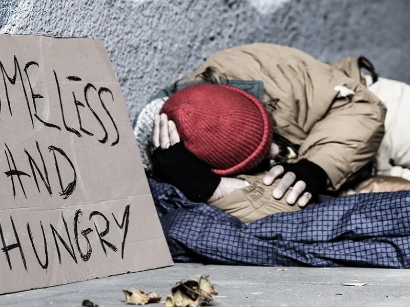 Photo of a homeless person sleeping outdoors on a sidewalk, next to a sign that says "homeless and hungry." Photo by Photographee.eu on Adobe Stock.